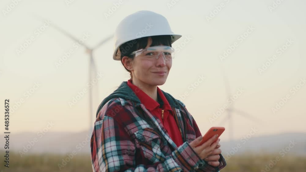 Portrait engineer woman in hard hat and goggles stand use phone look at camera smiling on ecological station. Wind turbines rotating. Slow motion Portrait engineer woman in hard hat and goggles stand use phone look at camera smiling on ecological station. Wind turbines rotating. Slow motion