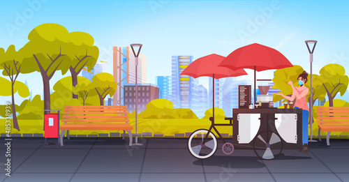 seller in mask standing near small street food market with cupcakes and coffee in urban park cityscape background