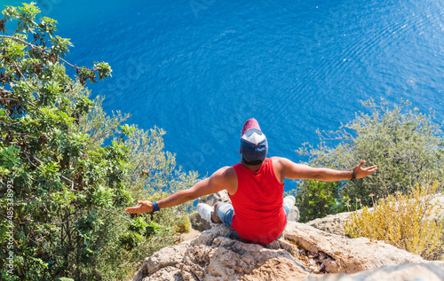 Turkish man sitting at the edge of a cliff with open arms and enjoying the beautiful sea scenery. View of Butterfly Valley in Turkey. Freedom Concept. photo