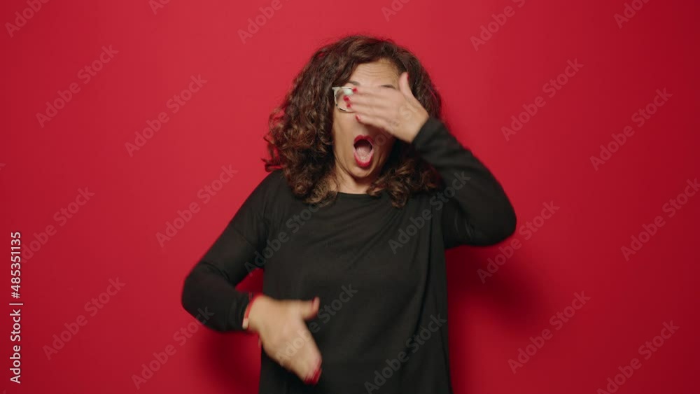 Middle age woman scared covering eyes with hand over red background Middle age woman scared covering eyes with hand over red background