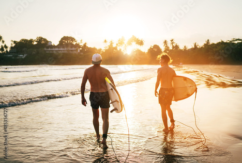 Photography Father with teenager son walking with surfboards by the sandy ocean beach with palm trees on background lightened with sunset sun