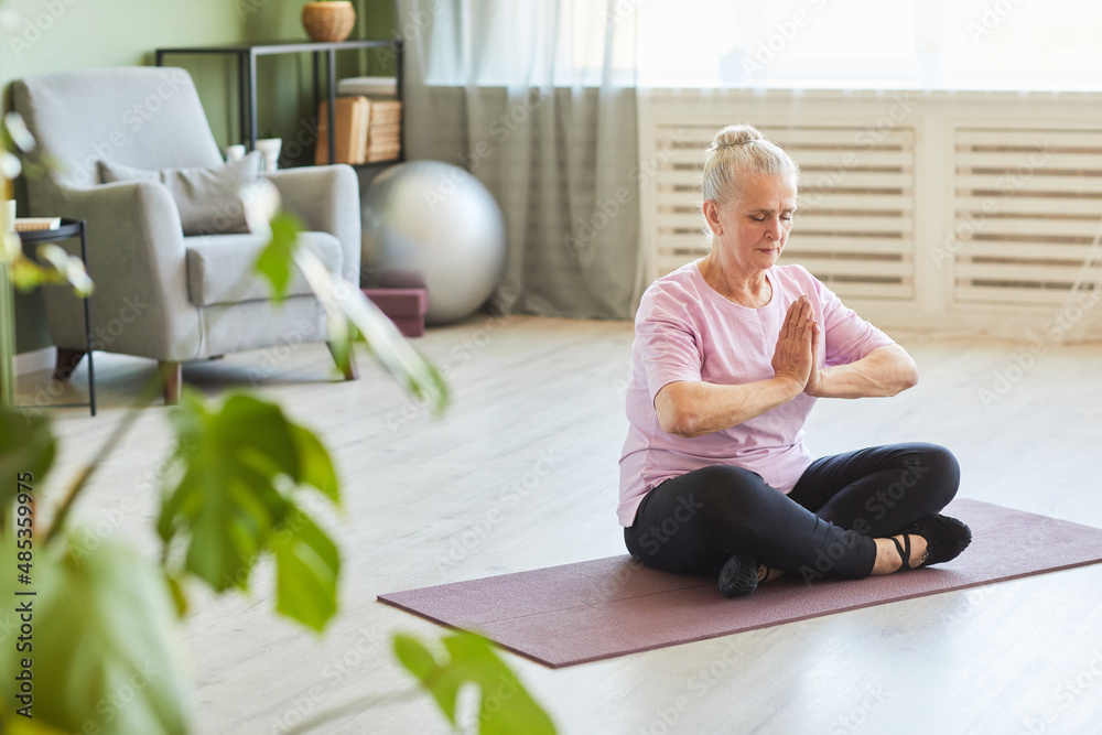 Serene senior woman in sports clothes sitting on mat and keeping hands put together by chest during yoga exercise