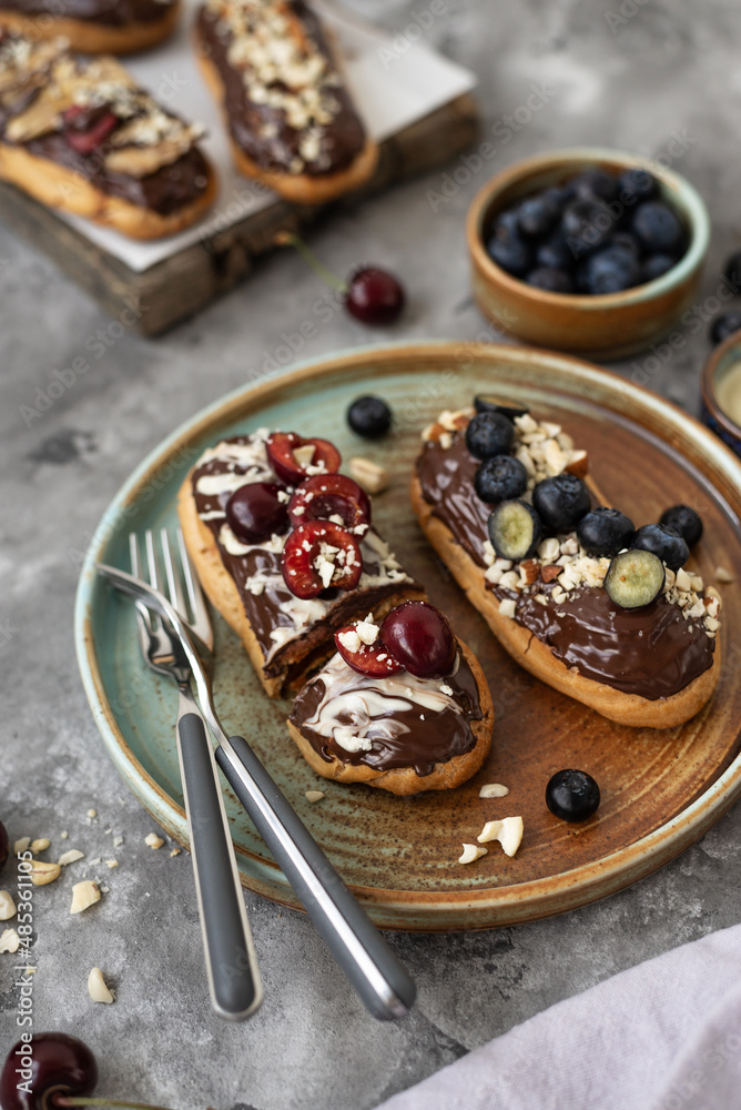 Eclairs with chocolate topping, nuts and berries. Chocolate, nut, banana, cherry and blueberry eclairs. Dessert on a plate. Top view Eclairs with chocolate topping, nuts and berries. Chocolate, nut, banana, cherry and blueberry eclairs. Dessert on a plate. Top view