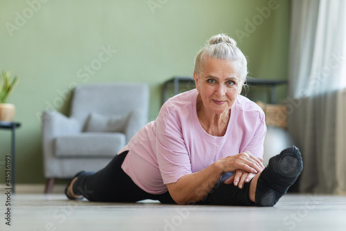 Senior active woman in sports clothes looking at camera while sitting in yoga position called hanumanasana on the floor of living-room photo