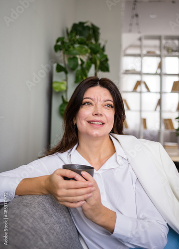 a happy brunette woman in a suit is sitting in an armchair with a cup of coffee. Break in work