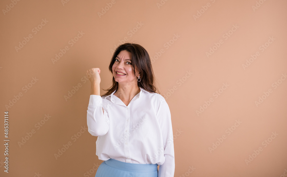 a satisfied brunette woman in a white shirt stands and shows success with her hand on a beige background with a place for text