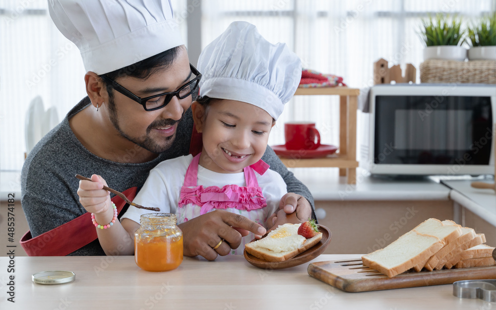 Asian young father with beard and little mixed race cute daughter with apron and chef hat smiling and preparing breakfast like spread jam on bread with strawberry together in kitchen at home Asian young father with beard and little mixed race cute daughter with apron and chef hat smiling and preparing breakfast like spread jam on bread with strawberry together in kitchen at home