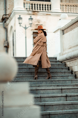 Outdoor fashion portrait of young elegant fashionable brunette woman, model in stylish hat, choker and light raincoat posing at sunset in European city.