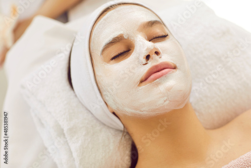 Beautiful young woman in beauty salon does skin care treatments and enjoys moisturizing face mask. Close up of woman with white headband lying with her eyes closed on massage table. Beauty concept.