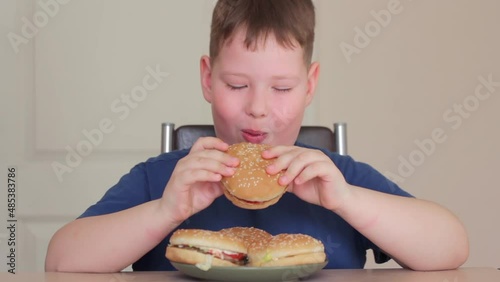 A fat boy eats a hamburger. He is sitting on a chair and very happy.
