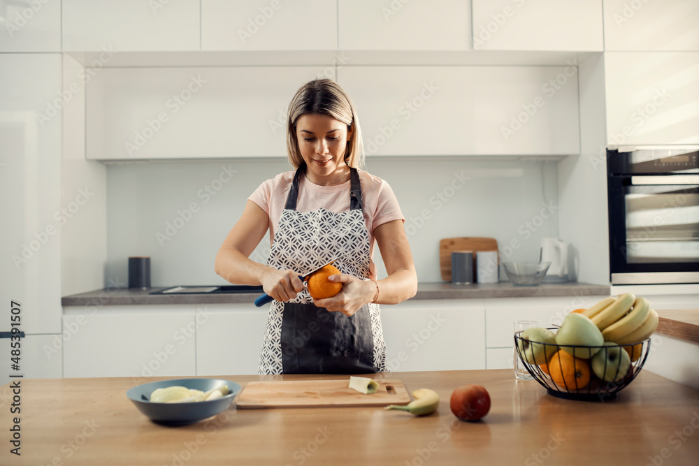 A happy blonde cutting orange and preparing healthy snack while standing in her modern kitchen. A happy blonde cutting orange and preparing healthy snack while standing in her modern kitchen.