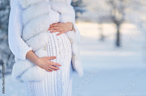 A pregnant woman in a white knitted dress and a fur vest in the forest in winter.