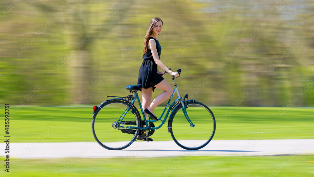 a young woman in a black short dress quickly rides a bicycle on a summer green park