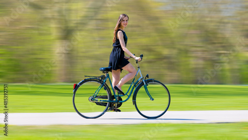 a young woman in a black short dress quickly rides a bicycle on a summer green park