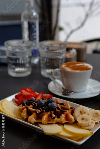 Traditional Italian breakfast. Fresh classic waffles with fruits, sugar powder, chocolate and cappuccino coffee with cinnamon in Milan, Lombardy, Italy. European food, drinks  pastry at a local bar.