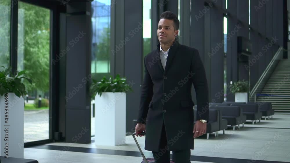 Young male executive moving suitcase and walking inside office or airport building spbd. Close-up view of American African man holds baggage in hand and looks with smile, walks in comfortable lobby Young male executive moving suitcase and walking inside office or airport building spbd. Close-up view of American African man holds baggage in hand and looks with smile, walks in comfortable lobby