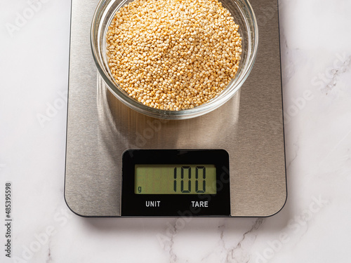 Raw dry quinoa in a glass bowl on a digital kitchen scale. 100g white quinoa seeds on the scales displaying grams. Rectangular electronic device for food measurement during dieting and cooking. photo