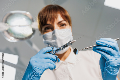 Woman dentist in blue face mask and gloves examine teeth with syringe and mirror. View from patient seat to doctor. Creative photo on dental theme. Concept of tooth treatment without pain in clinic.