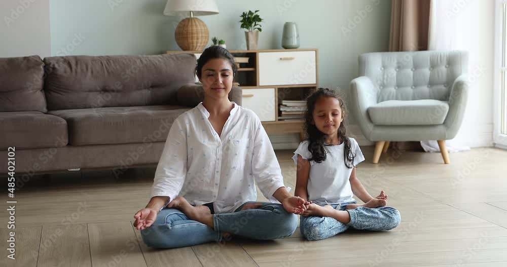 Attractive Indian yogini mom teach good life habits her cute little 6s daughter, sit on warm floor cross-legged, do meditation practice together in living room. Yoga at home, healthy lifestyle concept Attractive Indian yogini mom teach good life habits her cute little 6s daughter, sit on warm floor cross-legged, do meditation practice together in living room. Yoga at home, healthy lifestyle concept