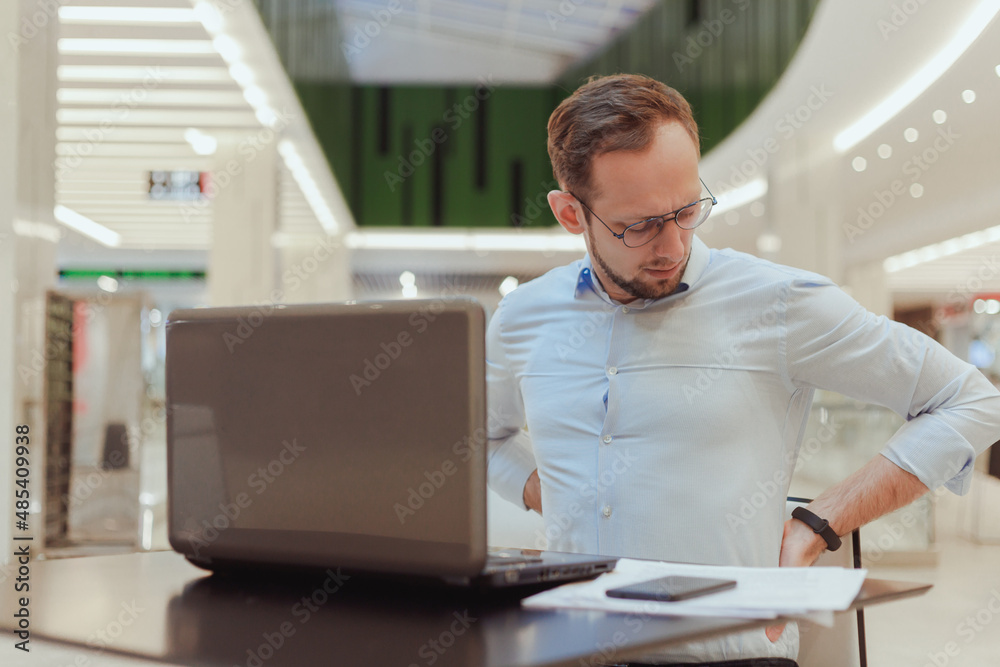 Fototapeta premium Man holds on to his back, suffers from lower back pain, working on computer. Sedentary work office worker
