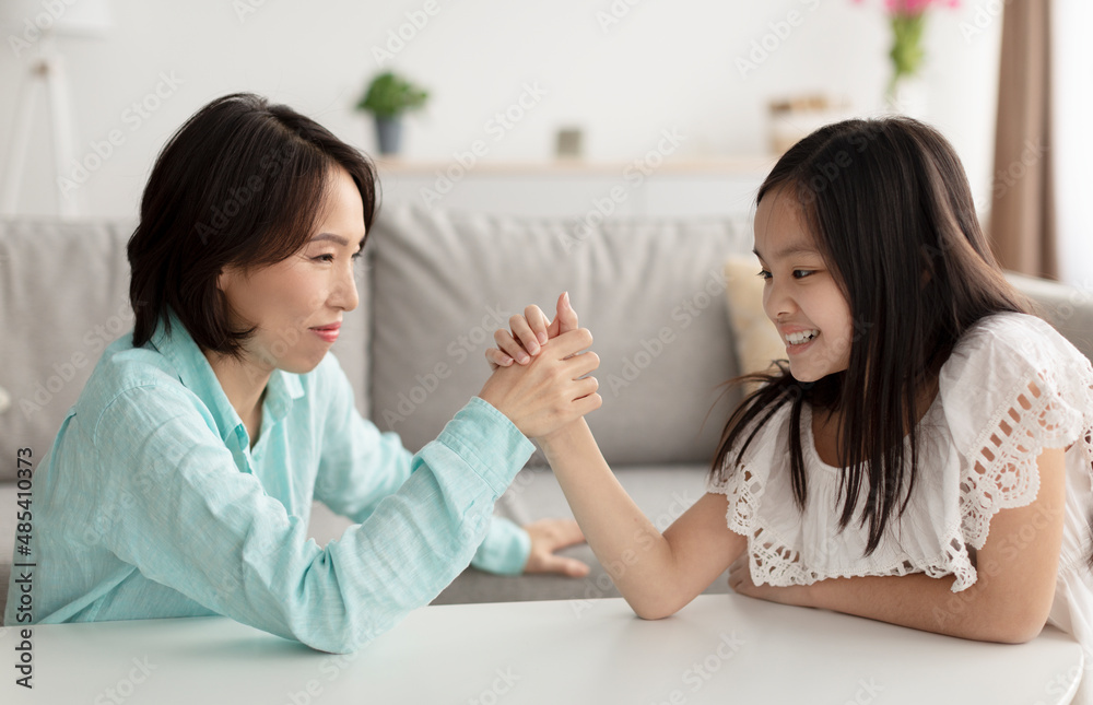 Stressed little Asian girl arm wrestling with her grandmother, having conflict, fighting at home