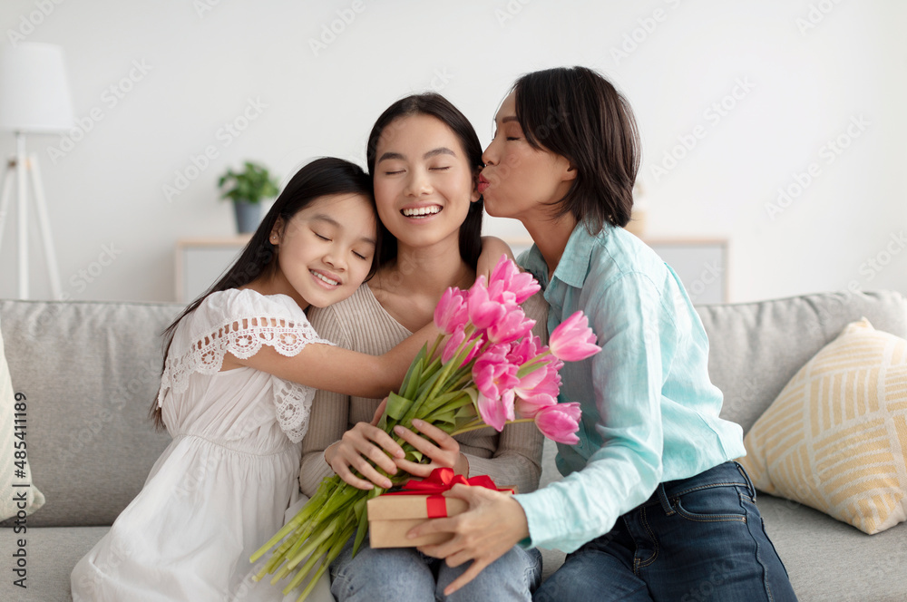 Young Asian woman with flowers and gift box being kissed and hugged by her mature mom and cute little daughter at home