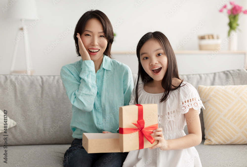Excited mature Asian woman and her adorable little granddaughter opening gift on couch at home Excited mature Asian woman and her adorable little granddaughter opening gift on couch at home