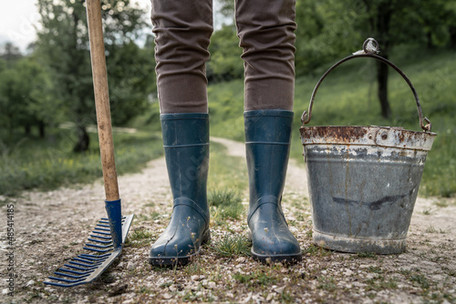 Close up on legs of unknown woman in rubber boots standing on the dirt road with metal bucket and rake beside - farm agriculture and sustainable life concept © Miljan Živković