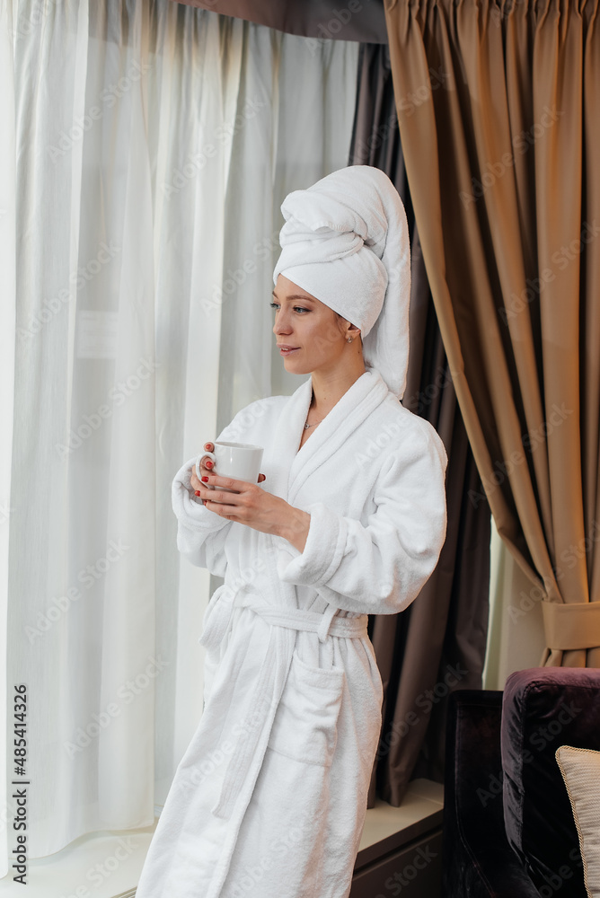 A young beautiful girl in a white coat is standing and drinking coffee near the window in her hotel room. Rest and travel. Hotel recreation, and tourism.