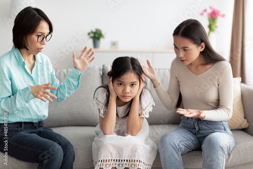 Murais de parede Unhappy Asian girl covering ears while angry mother and grandmother scolding her at home
