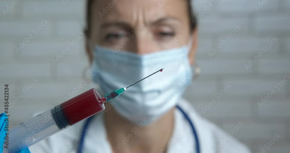 Hospital work with blood test. A view of doctor in protective mask with a syringe with blood. A concept of medical test during epidemic time. Hospital work with blood test. A view of doctor in protective mask with a syringe with blood. A concept of medical test during epidemic time.