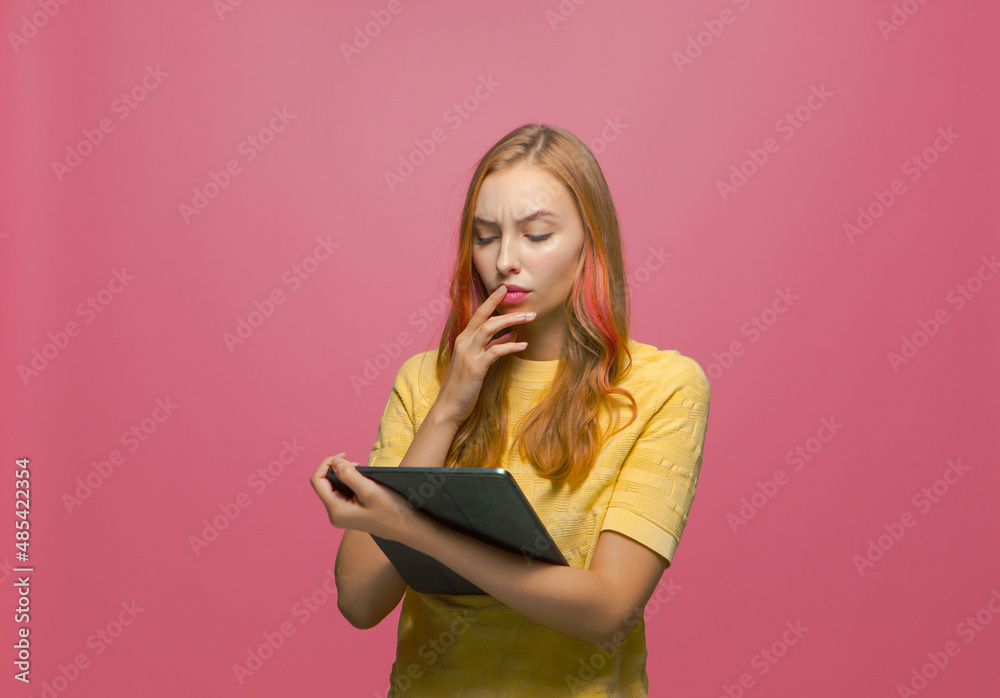 Pensive puzzled young girl looking at tablet, solving problem, thinking out idea, making choice on pink background Pensive puzzled young girl looking at tablet, solving problem, thinking out idea, making choice on pink background