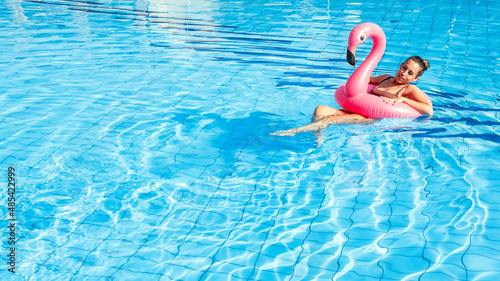 Summer time. Happy young sexy girl in bikini swimsuit with pink inflatable flamingo relaxing in blue pool water. Swimming pool water, having fun and enjoying travel vacation. © Maksym