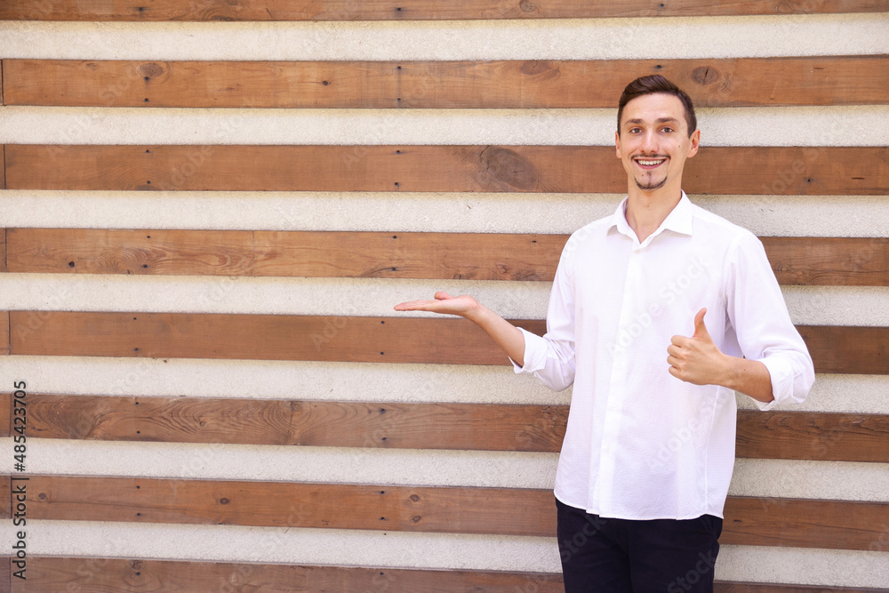 A happy Caucasian bearded man in a white shirt and black pants holding something in his palm and showing his thumbs up on a brown ribbed background with copy space A happy Caucasian bearded man in a white shirt and black pants holding something in his palm and showing his thumbs up on a brown ribbed background with copy space