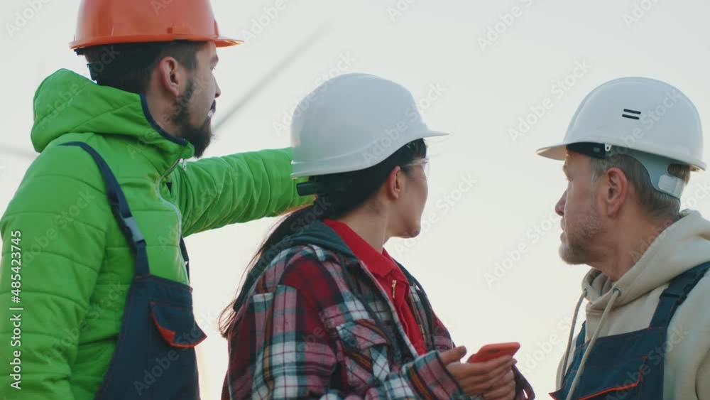 Three engineers in hard hat use mobile phone. On background of wind turbines renewable energetics. On ecological station at sunset. Slow motion Three engineers in hard hat use mobile phone. On background of wind turbines renewable energetics. On ecological station at sunset. Slow motion