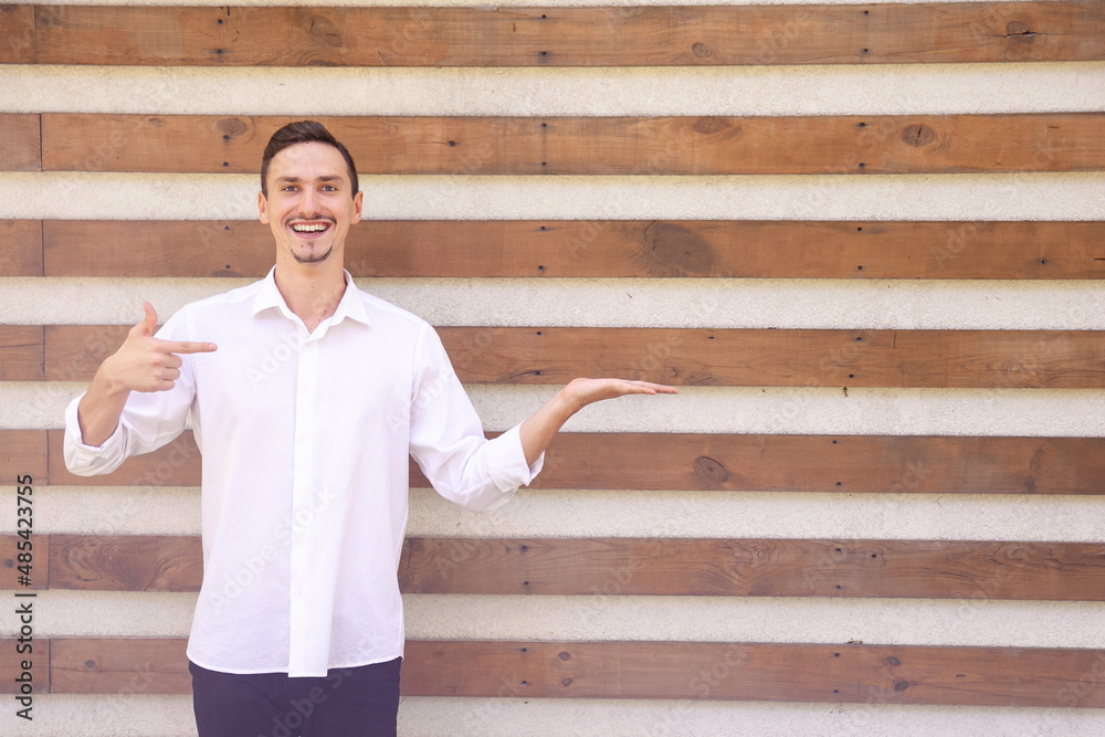 Obraz premium a happy European bearded guy in a white shirt holding something in the palm of his hand and pointing his finger at it and on a brown ribbed background with copy space