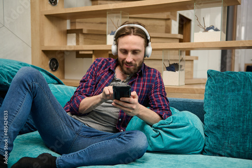 Caucasian young man using social media at home. Handsome guy with wireless headphones relaxing on the couch at home © Taras Grebinets