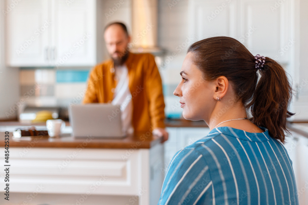 Upset wife or Girlfriend looking towards husband after conflict argument While man use laptop.Young married couple after quarrel fight in kitchen home interior. Negative emotions in bad relationships. Upset wife or Girlfriend looking towards husband after conflict argument While man use laptop.Young married couple after quarrel fight in kitchen home interior. Negative emotions in bad relationships.