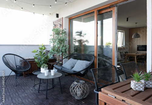 Beautiful terrace with rattan and wooden furniture, chair, table and sofa with pillow. Large window and lamps outdoor. Modern patio with plants, stylish design for relax end rest time. photo