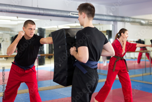 Caucasian man exercising jabs with teenage boy who holding punching pad during group karate training.