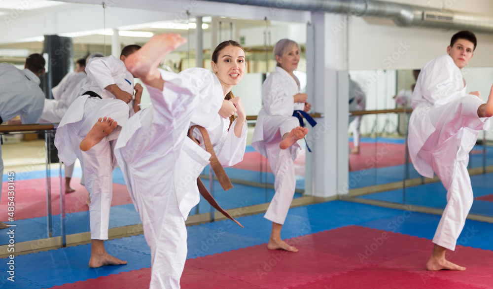 Athletic young woman in white kimono practicing leg kicks during group martial arts training at gym..