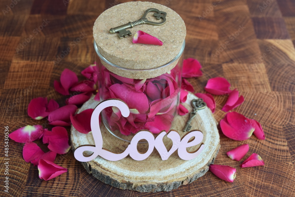 Vintage keys on top of glass container filled with rose petals on wooden round with the word love surrounded by rose petals. 