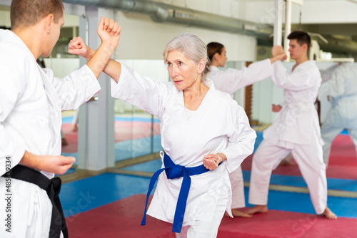 Sporty determined elderly woman in kimono practicing martial arts techniques paired with experienced trainer in training room