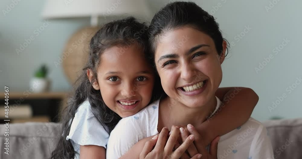 Little 6s Indian daughter piggyback her beautiful laughing mother, cheerful family hugging smiling looking at camera seated on sofa at home. Happy woman and child portrait, cherish and love concept