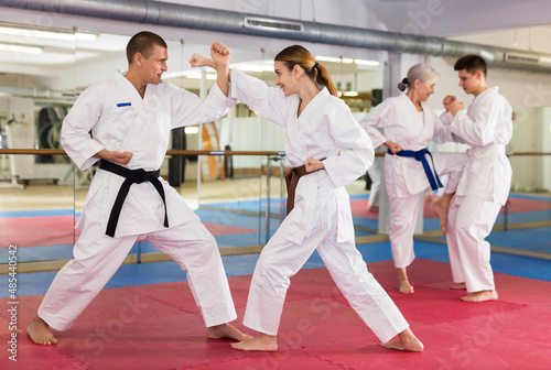 Woman and man in white kimono and belts sparring during karate training. Elderly woman and young man sparring in background.