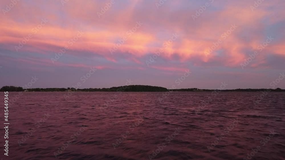 Aerial view of drone flying just over the surface of the water at sunset showing pink clouds and smaller white clouds with a forest in the background. drone flying over a lake Aerial view of drone flying just over the surface of the water at sunset showing pink clouds and smaller white clouds with a forest in the background. drone flying over a lake