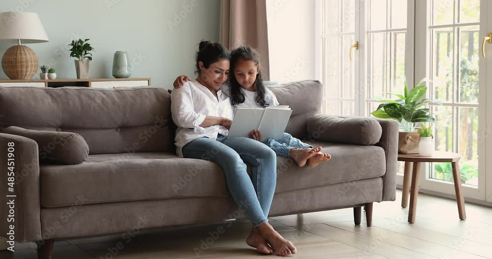 In cozy living room young Indian woman read book to her little 6s daughter, family sit on sofa spend weekend together enjoy favourite hobby. Storytelling, child development, leisure at home concept