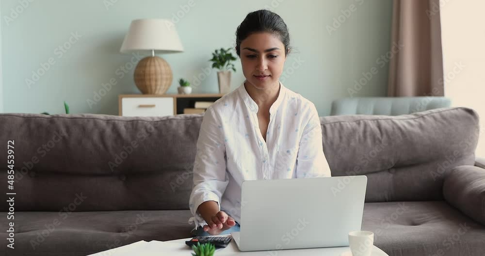 Serious Indian woman sit on sofa at table calculates expenses, pay utility bills use e-bank on laptop, sorts out paper, check documents, manage budget, control outcomes and incomes, accounting concept
