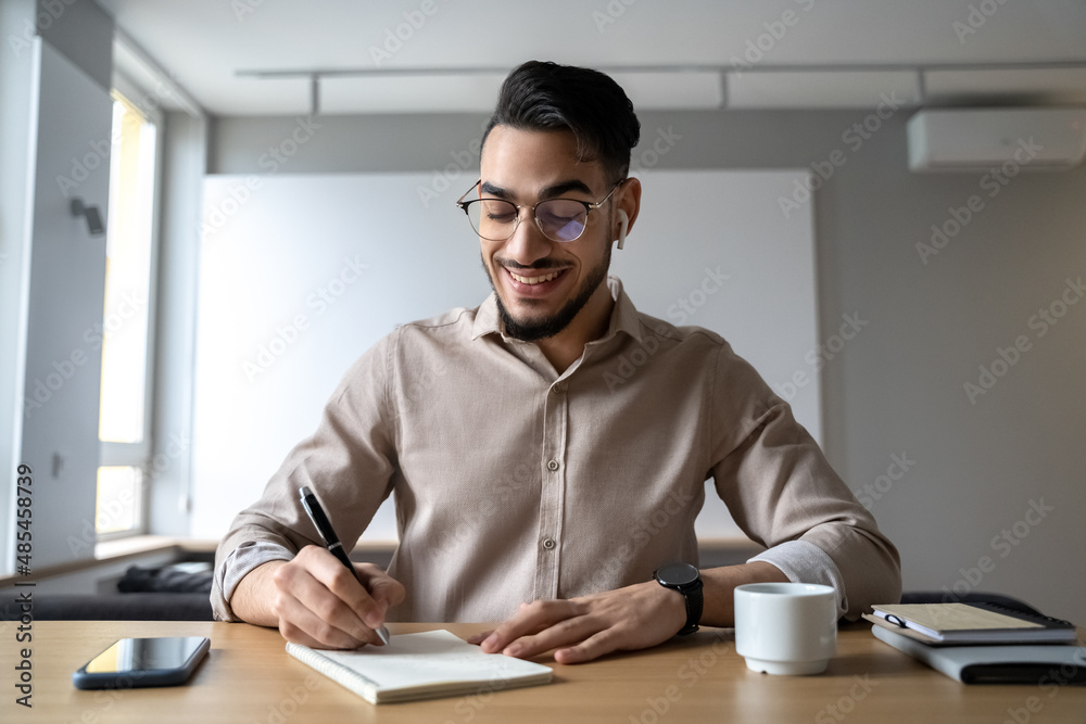 Happy smiling man sitting at desk writing in notepad listen online course Happy smiling man sitting at desk writing in notepad listen online course