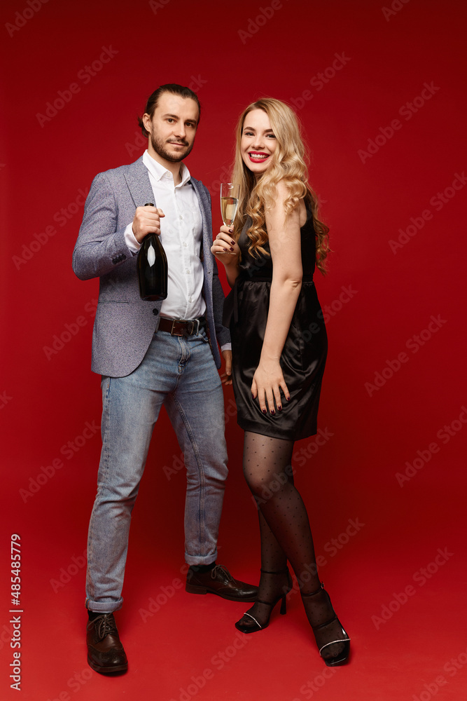 A handsome stylish man keeps a champagne bottle and a happy young woman in a black dress with champagne glass posing on the red background. Concept of 14th February mockup - st. Valentines day A handsome stylish man keeps a champagne bottle and a happy young woman in a black dress with champagne glass posing on the red background. Concept of 14th February mockup - st. Valentines day